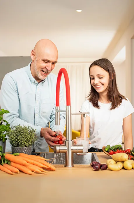 Ein Mann und ein Mädchen waschen Gemüse in einer modernen Küche. Sie lachen und teilen einen entspannten Moment. Vordergrund mit frischem Gemüse.