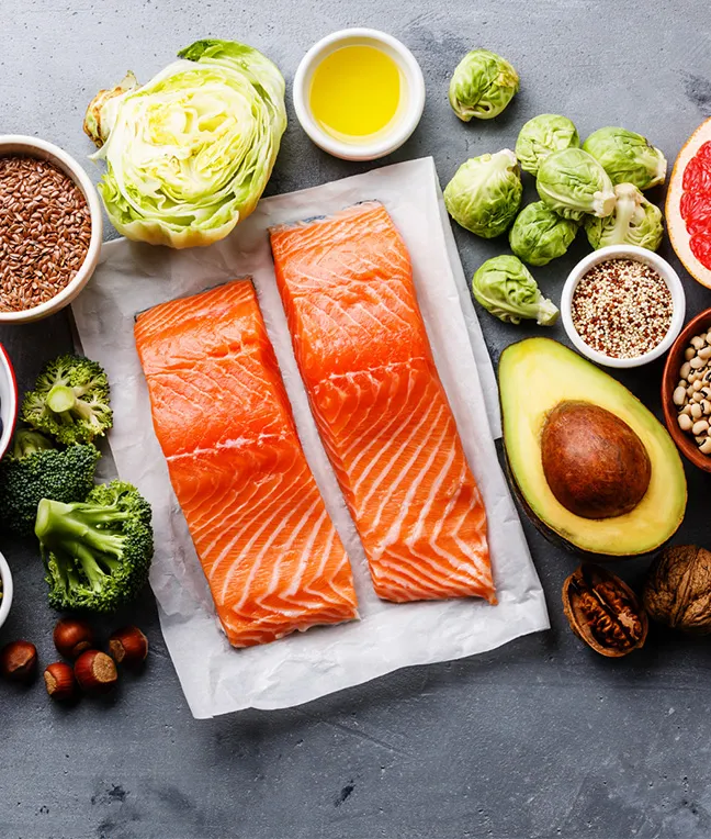 Zwei Lachsfilets auf Papier, umgeben von Gemüse wie Brokkoli, Rosenkohl und Salat, sowie Quinoa, Nüssen und Olivenöl.