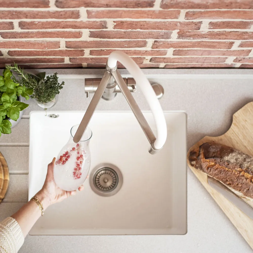 Eine Hand hält eine Glaskaraffe mit Wasser und Granatapfelkernen, während sie über einem eleganten Spülbecken in einer modernen Küche positioniert ist. Im Hintergrund ist eine Ziegelwand und frische Kräuter zu sehen.