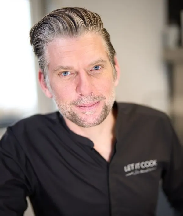 Porträt eines Mannes mit grauen Haaren, blauen Augen und Bart, der ein schwarzes Hemd trägt.