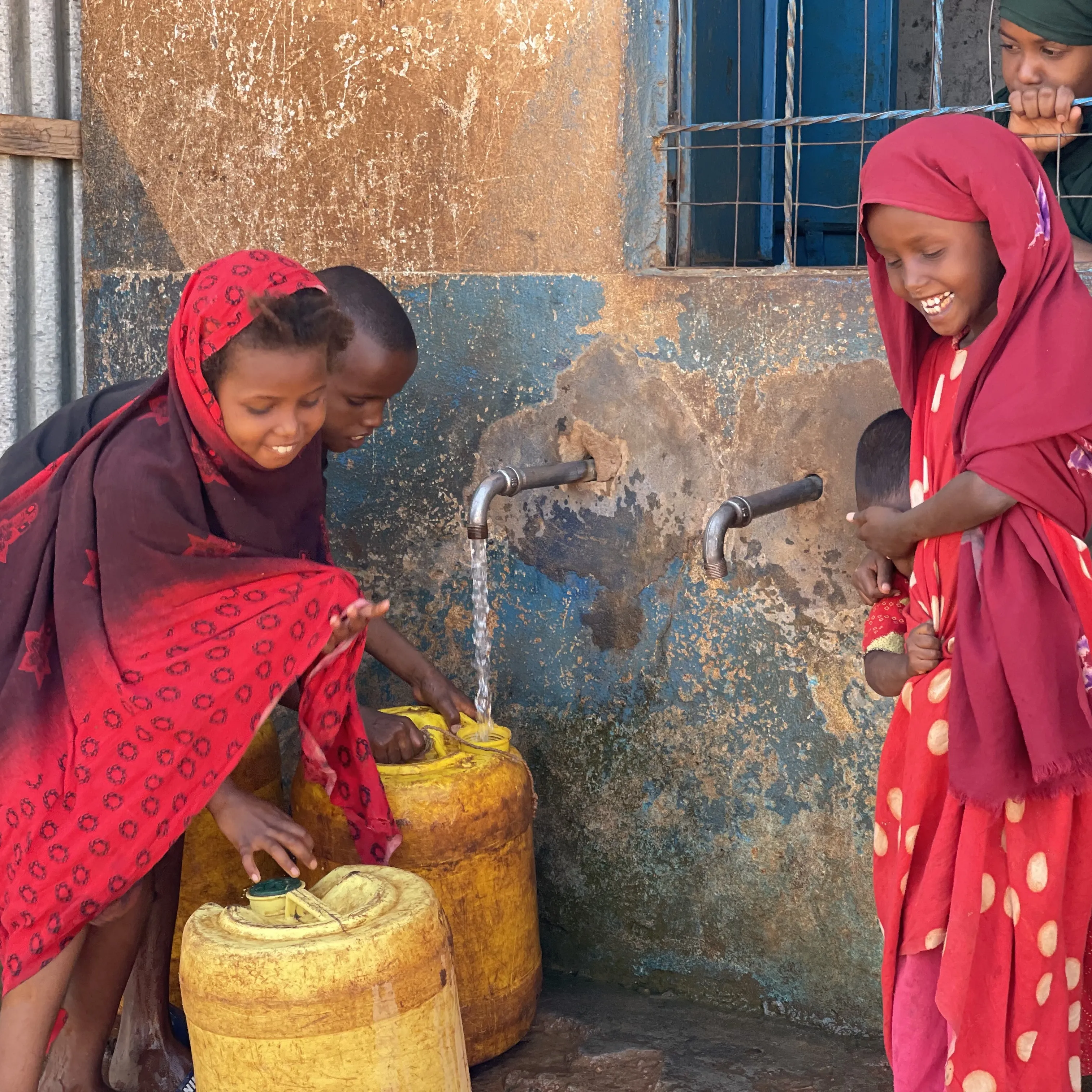 Drei Kinder füllen gelbe Wasserkanister an einem Wasserhahn und lächeln dabei.
