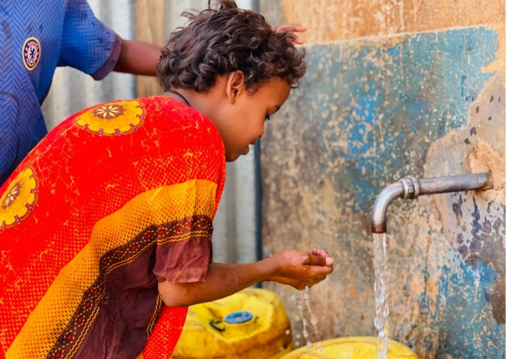 Ein Kind in einem bunten Shirt füllt mit Händen Wasser aus einem Wasserhahn in gelbe Kanister.