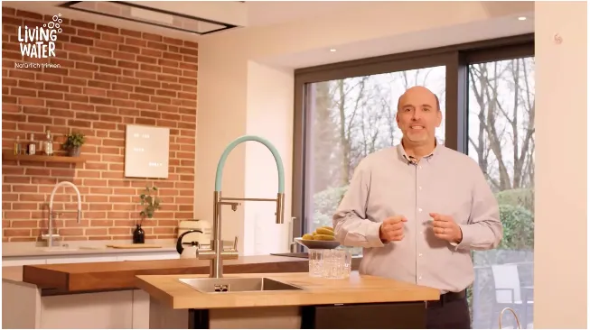 Ein Mann steht in einer modernen Küche mit einem Holzarbeitsplatz und einer Spüle. Im Hintergrund sind Pflanzen und Fenster zu sehen.