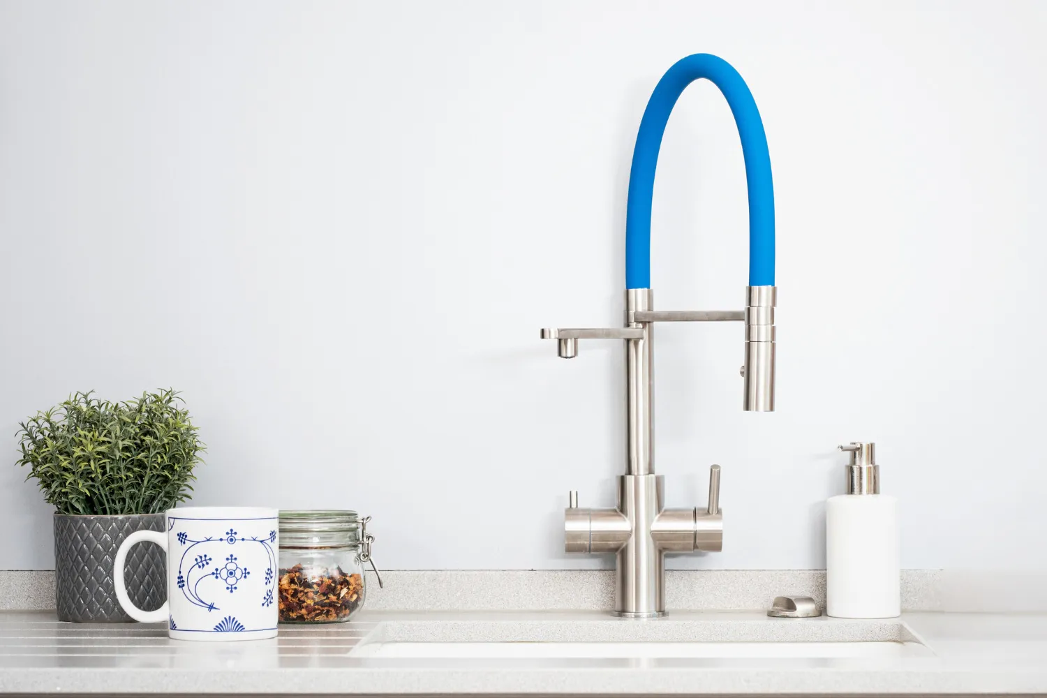 Moderne Küchenarmatur aus Edelstahl mit blauem Schlauch, einer Pflanze in einem Topf, einer Kaffeetasse und einem Glas mit Gewürzen auf einer Arbeitsfläche.