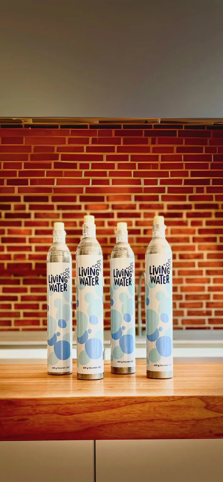 Vier Flaschen mit dem Aufdruck „Living Water“ auf einem Holztablett vor einer rotkarierten Wand.