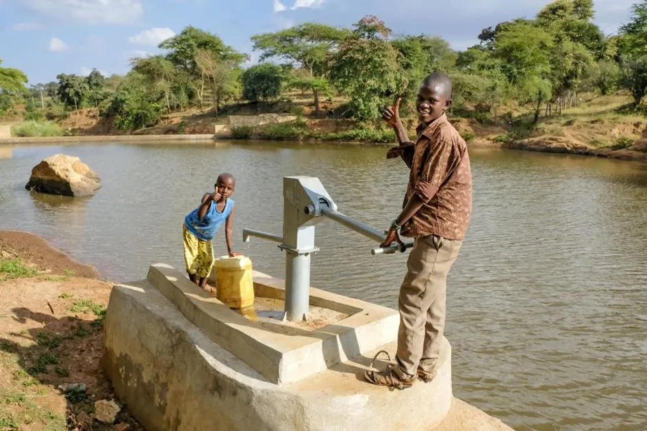 Eine Pumpe steht am Ufer eines Gewässers. Ein Junge steht neben einem Mann, der an der Pumpe arbeitet und beide lächeln.