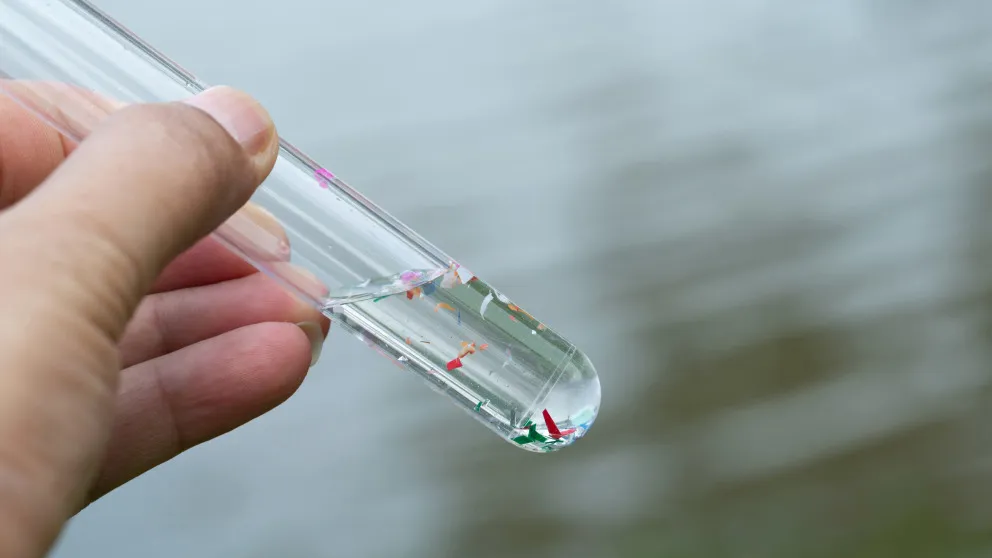 Hand hält ein Reagenzglas mit Wasserprobe, darin schwimmen bunte Mikroplastikpartikel.