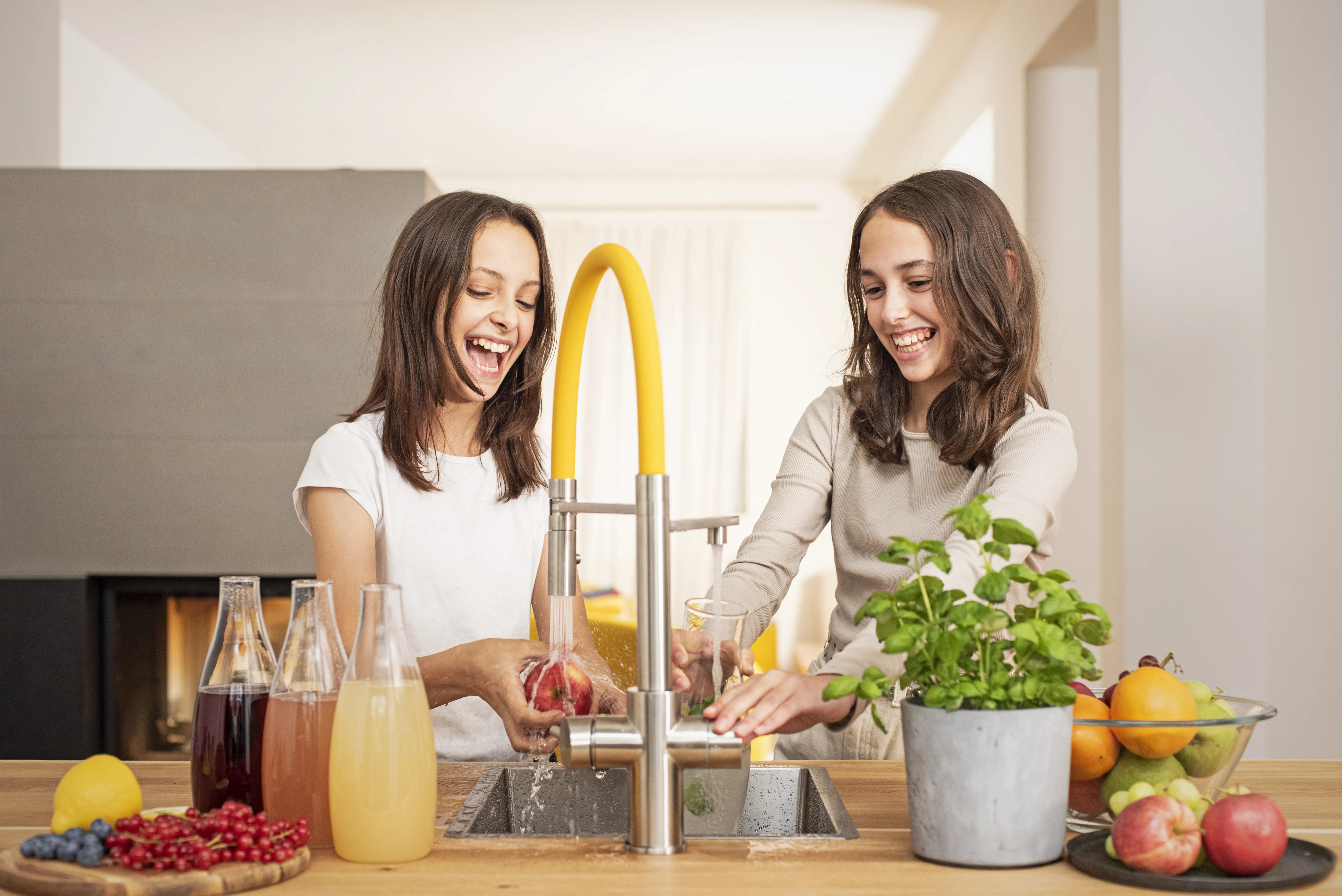 Zwei Mädchen lachen in der Küche und nutzen gemeinsam den Living Water Wasserhahn, daneben stehen Früchte und Karaffen.