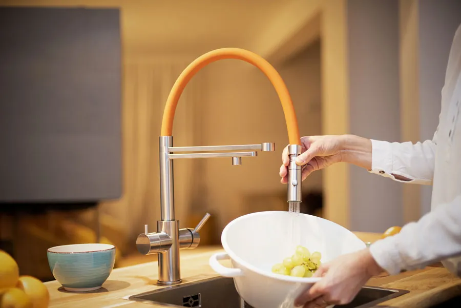 Moderne Living Water Küchenarmatur mit flexiblem orangefarbenem Schlauch – eine Person wäscht frische grüne Trauben in einer weißen Schüssel unter fließendem Wasser.