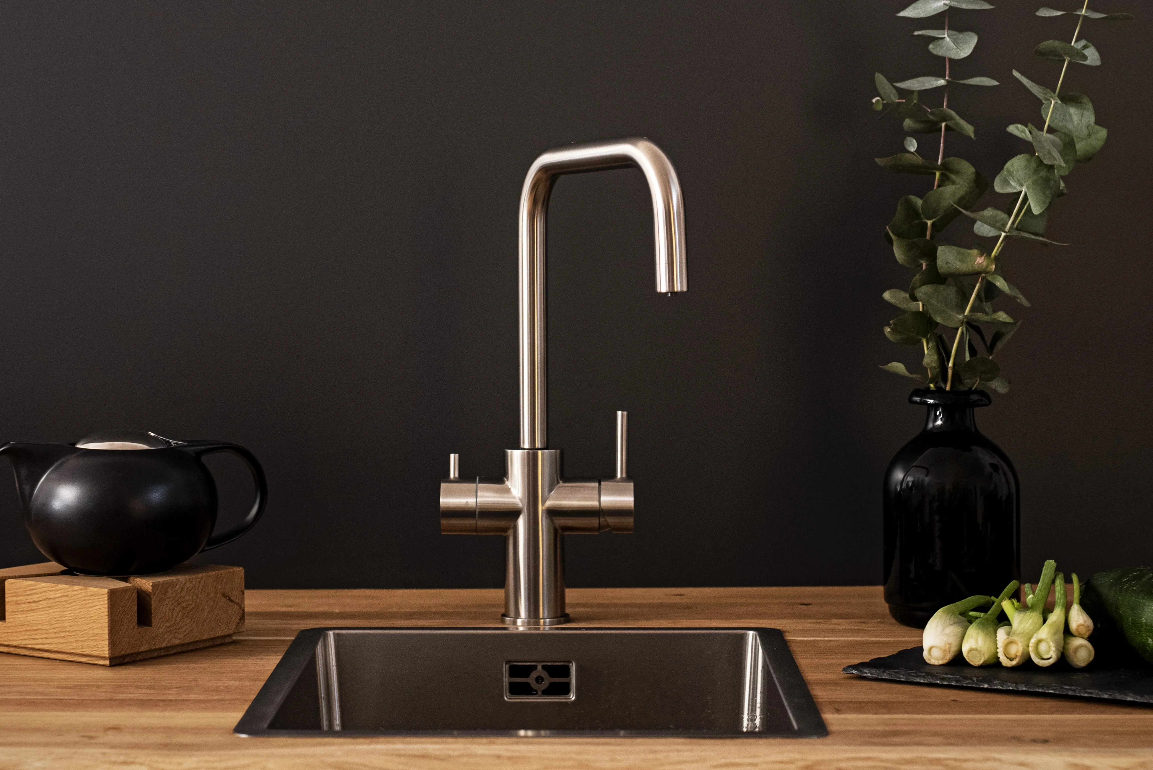 Stylische Küchenspüle mit Edelstahlarmatur, schwarzem Kännchen, Eukalyptuszweigen und Gemüse.