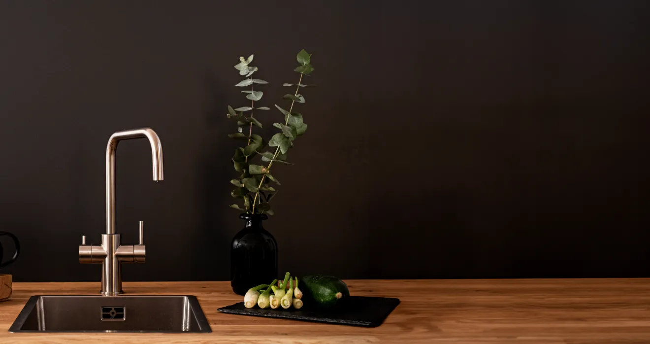 Eine moderne Küchenzeile mit einem Edelstahl-Wasserhahn, einer Spüle, einer schwarzen Vase mit Eukalyptus und frischem Gemüse auf einem schwarzen Brett.