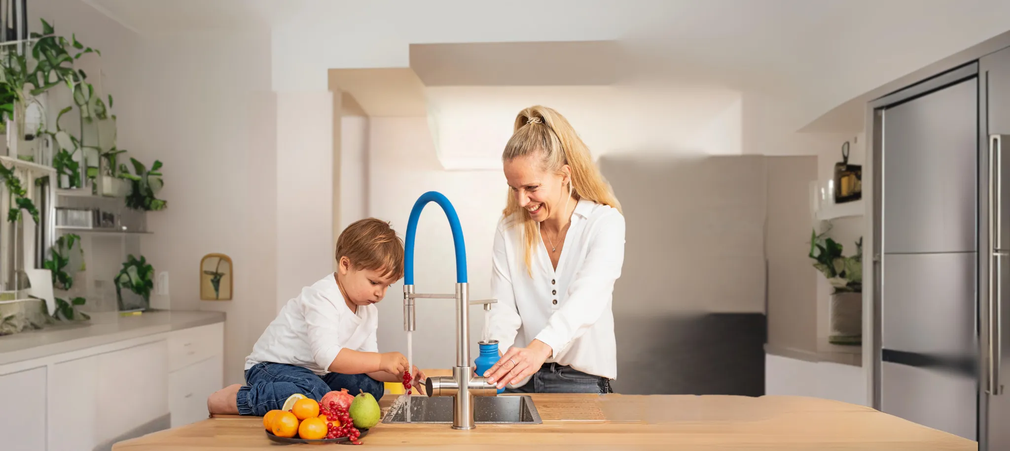 Eine Frau und ein Kind stehen an einer modernen Küchenarbeitsplatte mit einem Wasserhahn. Das Kind spielt mit Obst, während die Frau lächelt. Pflanzen sind im Hintergrund zu sehen.