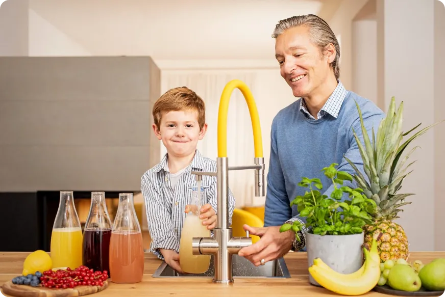Ein Junge und ein Mann stehen an einer Spüle und bereiten Getränke vor. Neben ihnen stehen Flaschen mit verschiedenen Säften und frisches Obst.