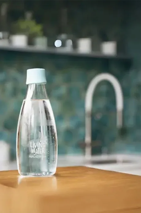 Eine Glasflasche mit Wasser auf einem Holzblock, im Hintergrund eine moderne Küche.