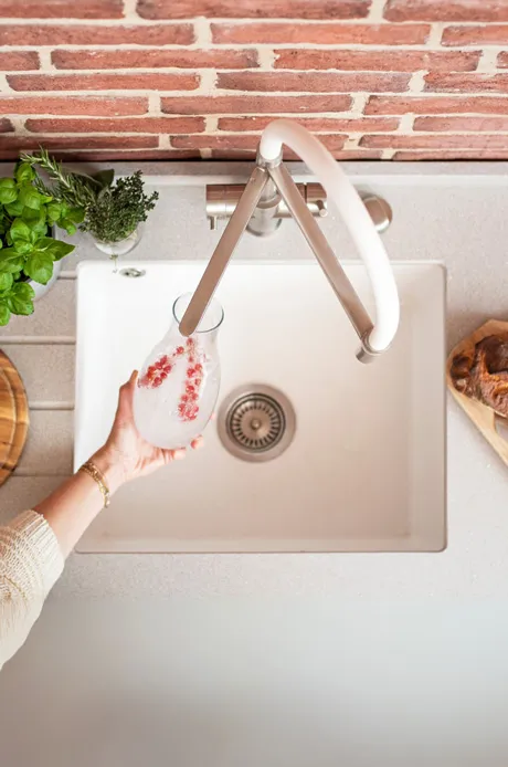 Eine Person hält ein Glas mit Getränk über einem Spülbecken, während frische Kräuter und eine Holzplatte im Hintergrund zu sehen sind.