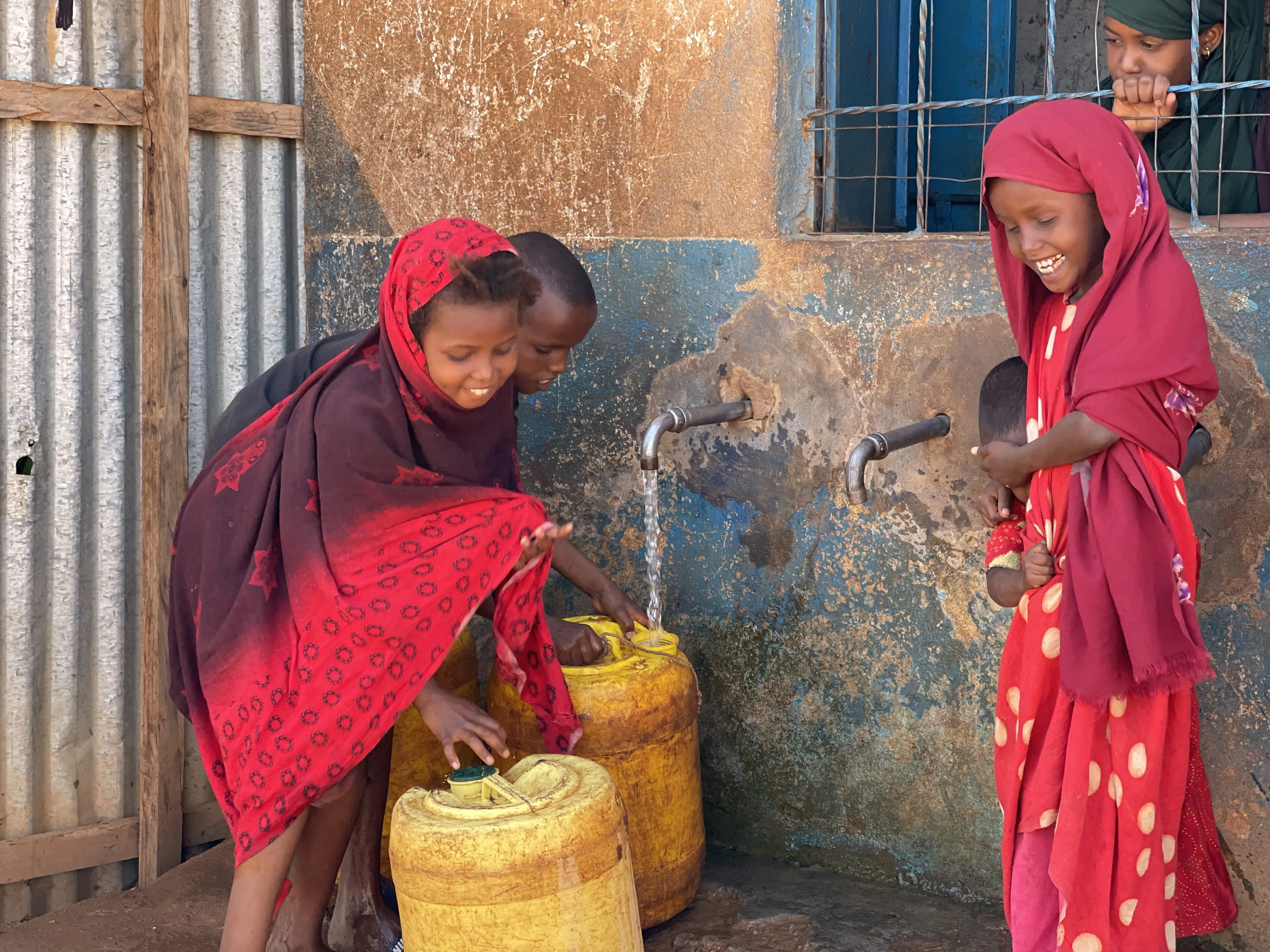 Kinder füllen Wasser aus einem Wasserhahn in gelbe Kanister. Sie tragen farbenfrohe Kleider und lächeln.