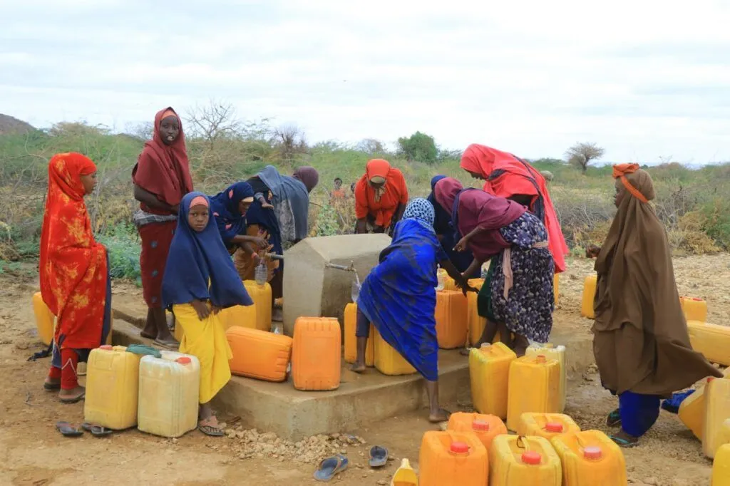 Frauen und Mädchen holen Wasser an einem Brunnen, umgeben von gelben Wasserkanistern in einer trockenen Landschaft.