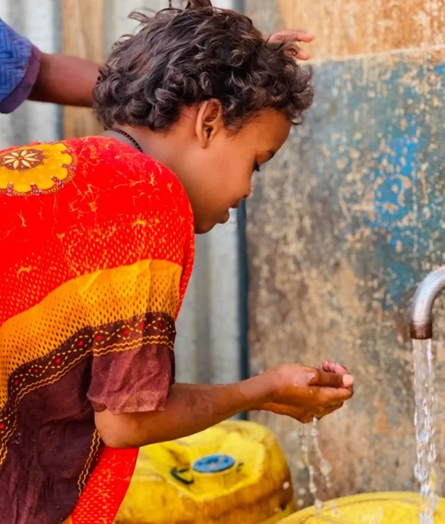 Ein Kind mit lockigem Haar in einem bunten T-Shirt fängt Wasser aus einem Wasserhahn in seinen Händen. Im Hintergrund sind gelbe Wassertanks sichtbar.