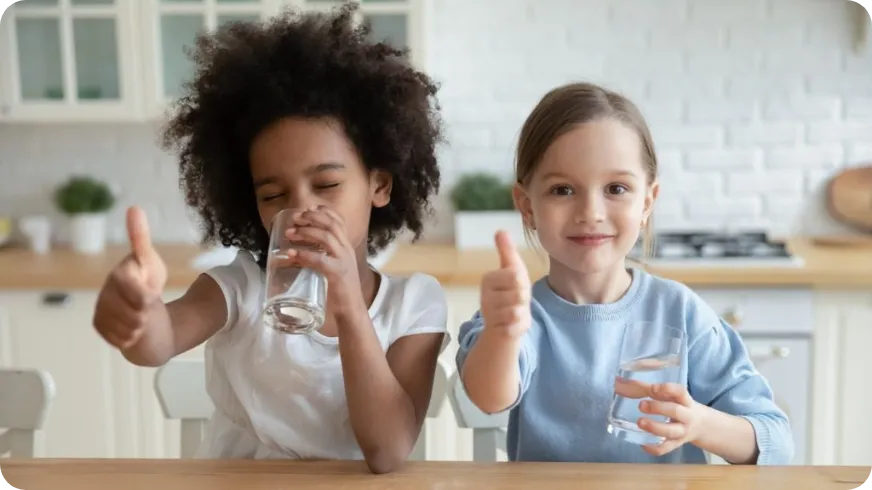 Zwei Kinder sitzen in der Küche und trinken Wasser, beide zeigen Daumen hoch.