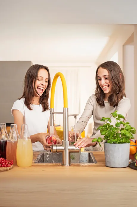 Zwei Mädchen stehen an einer Spüle und lachen, während sie mit einem Wasserhahn spielen. Im Hintergrund sind verschiedene Getränke und eine Pflanze sichtbar.