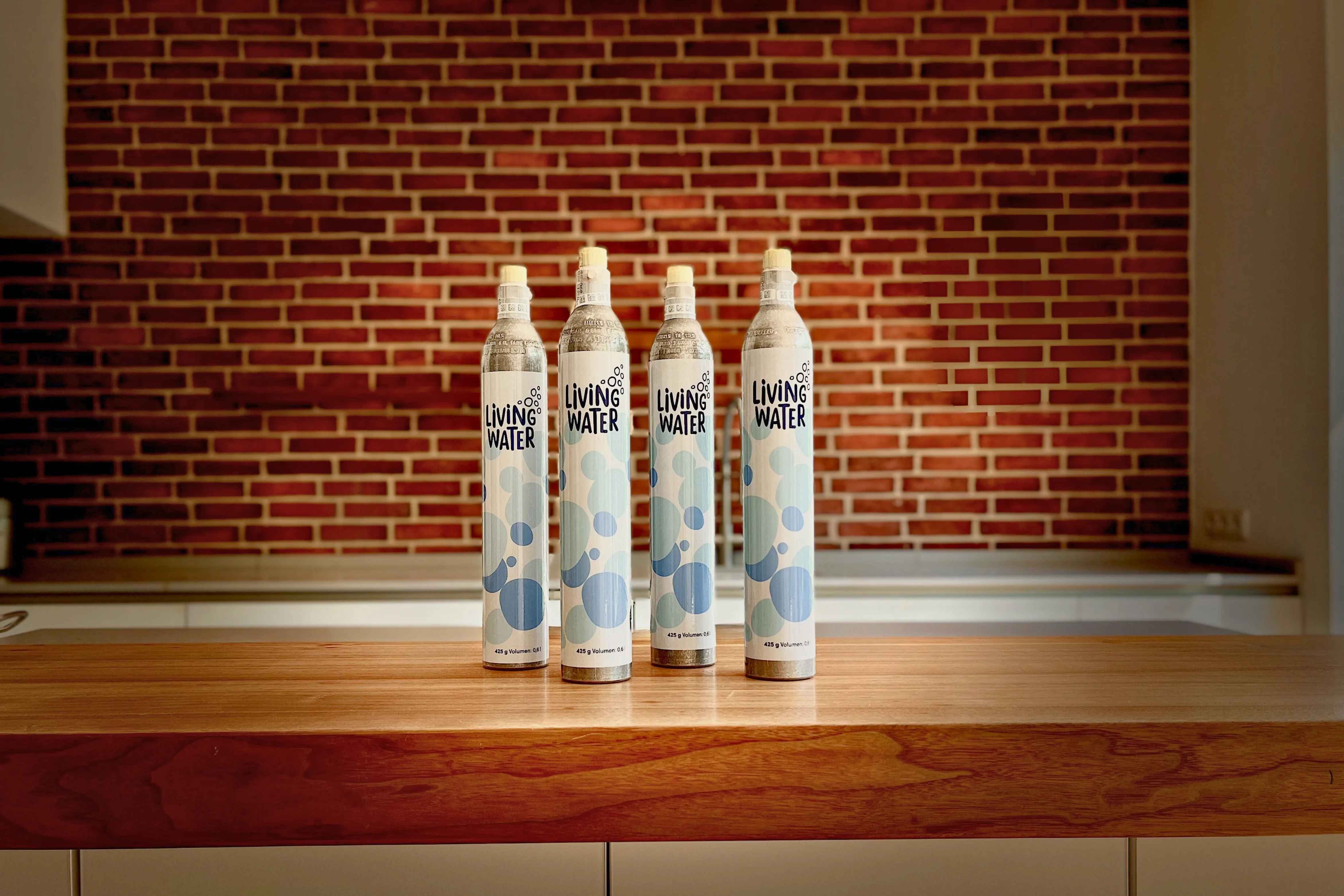 Vier Flaschen "Living Water" stehen auf einem Holzbrett vor einer Backsteinwand.