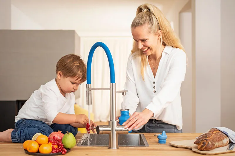 Eine Frau und ein Kind stehen an einer modernen Küchenarbeitsplatte mit einem Wasserhahn. Das Kind spielt mit Obst, während die Frau lächelt. Pflanzen sind im Hintergrund zu sehen.