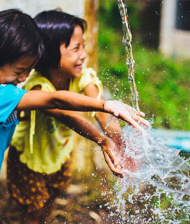 Zwei Kinder spielen lachend mit Wasser, das spritzt, während sie ihre Hände ausstrecken.