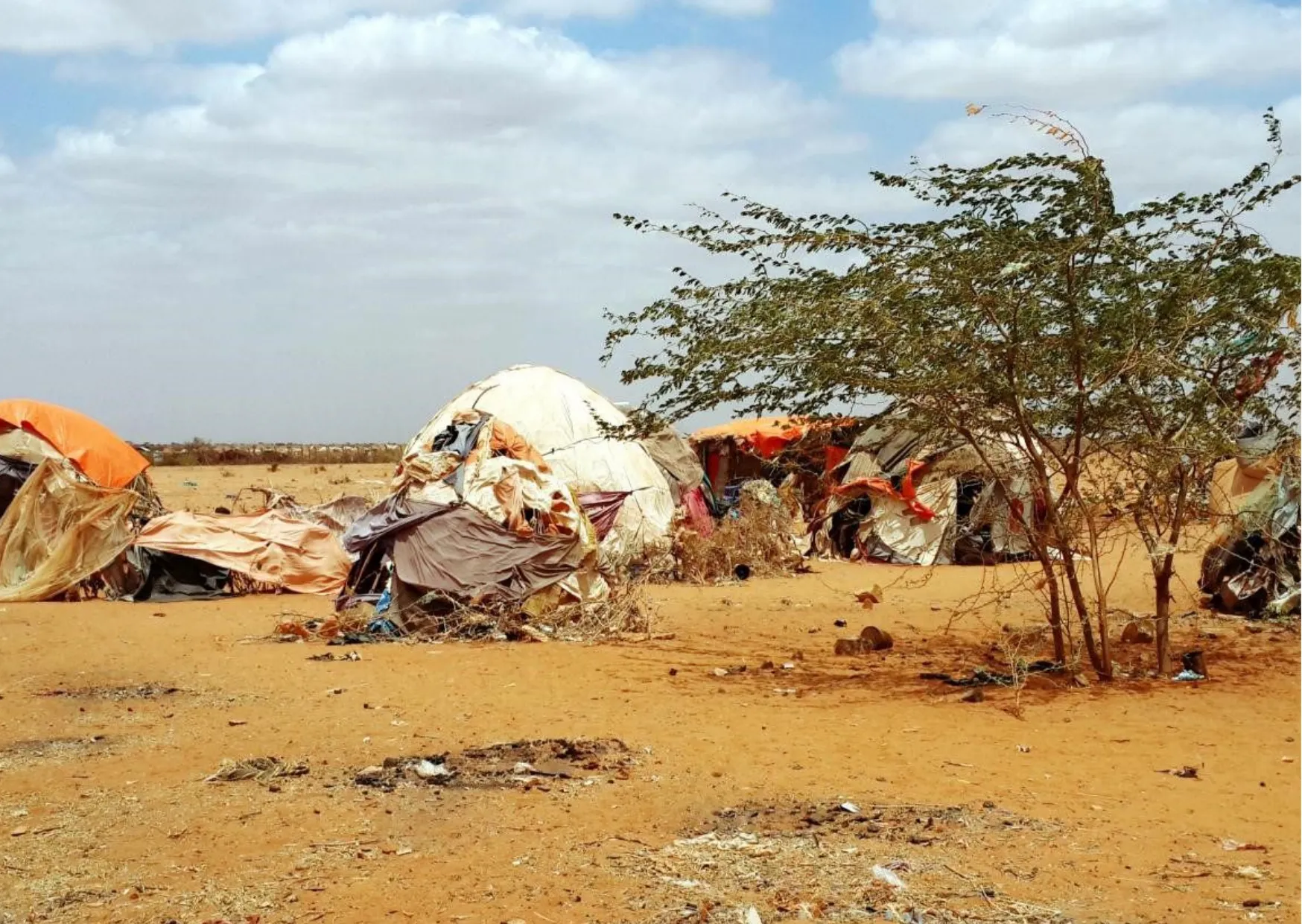 Zeltlager in einer Wüstenlandschaft mit beschädigten Überdachungen und spärlicher Vegetation.
