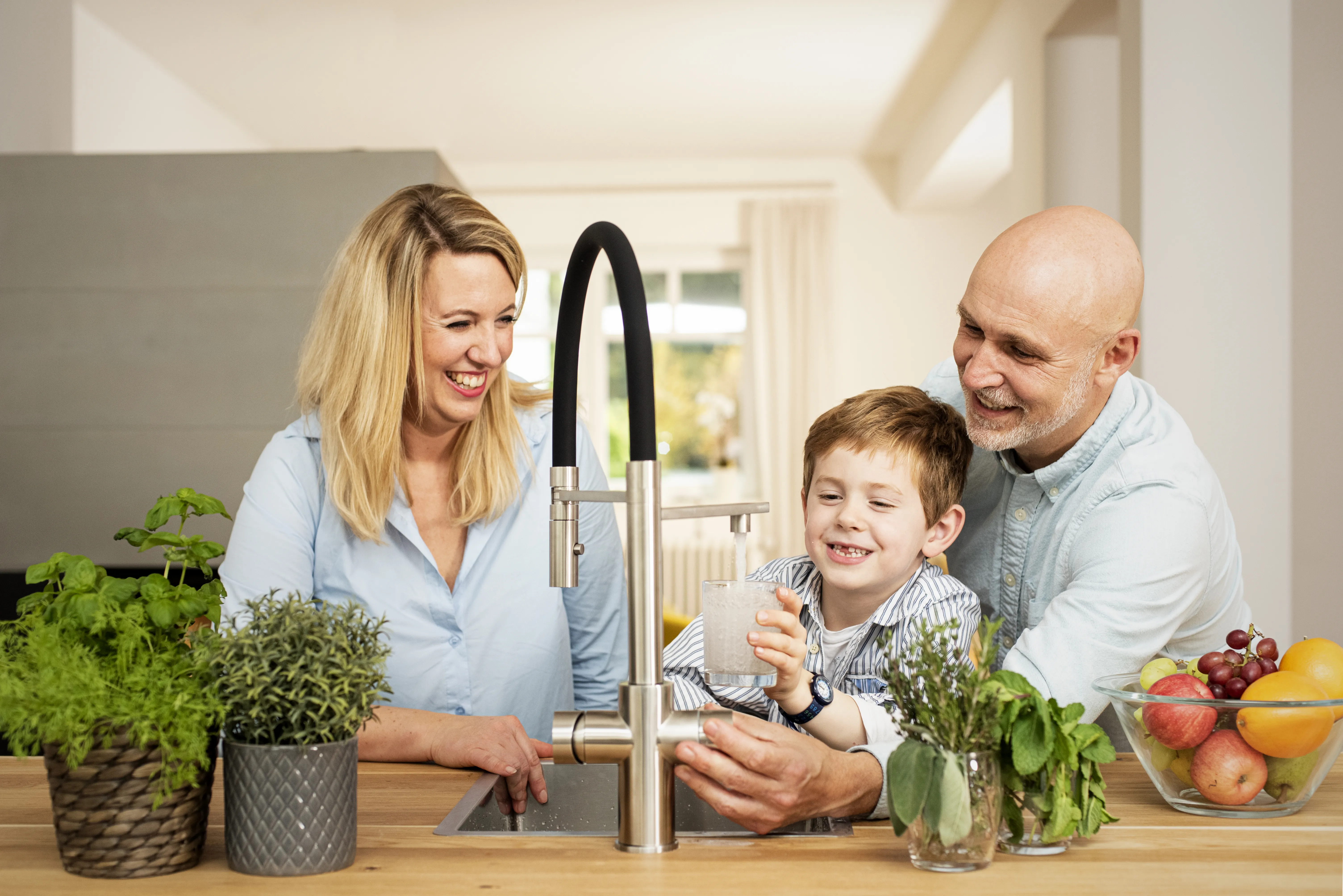 Ein fröhliches Familienmoment in der Küche: Eine Frau, ein Mann und ein Junge lächeln, während der Junge ein Glas mit Wasser füllt. Es gibt Kräuter in Töpfen und eine Schüssel mit Obst auf dem Tisch.