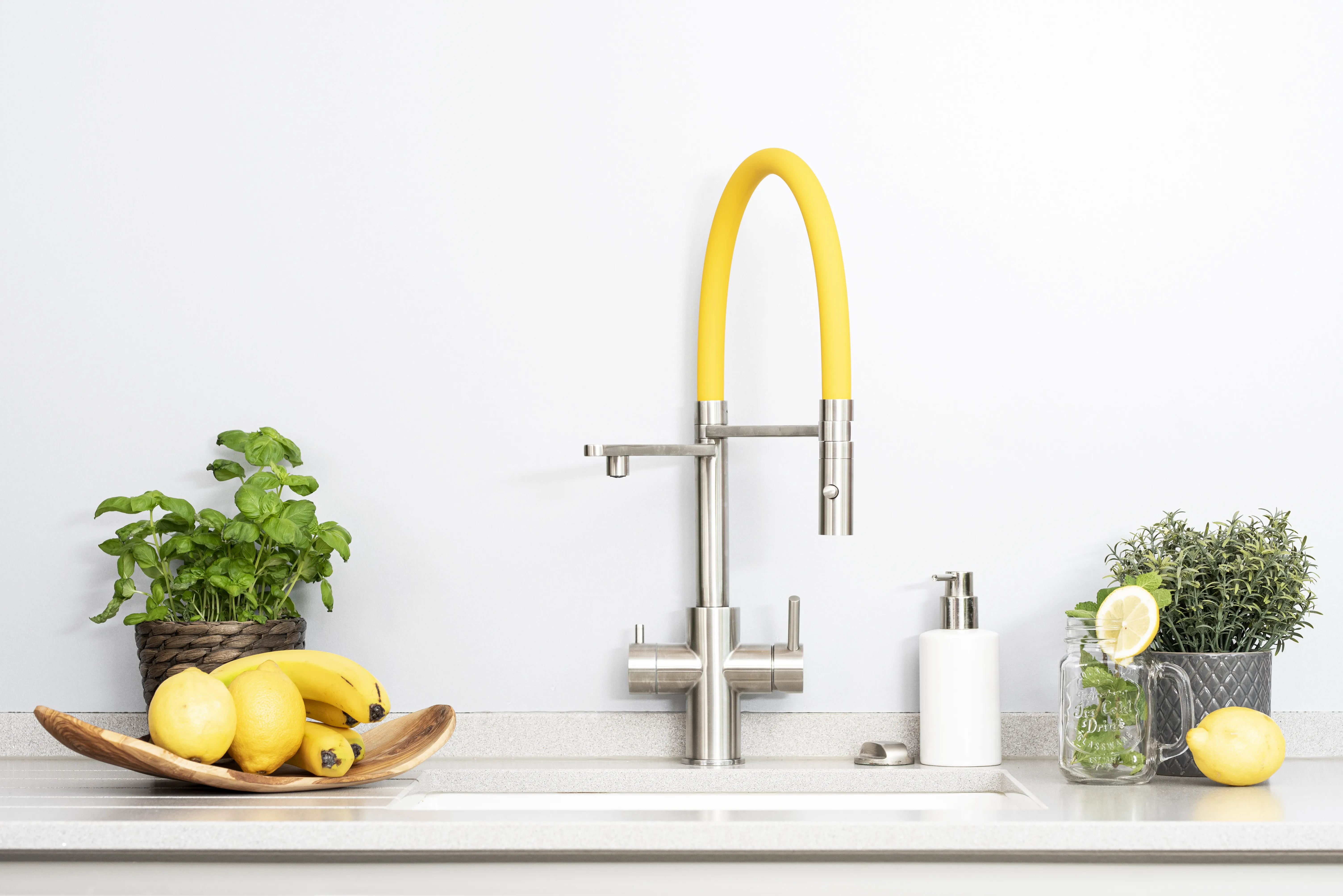 Eine moderne Küchenspüle mit einem Wasserhahn, der einen gelben Schlauch hat. Daneben befinden sich eine Holzschale mit Zitronen und Bananen sowie ein Glas mit einer Zitronenscheibe und Kräutern.